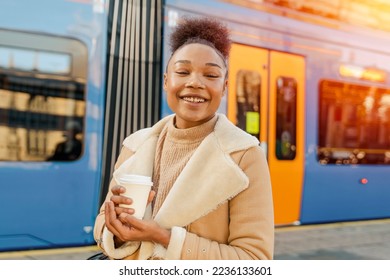 A cheerful woman holding a cup of coffee. A smiling curly brunette lady in a sweater waiting for a tram. - Shutterstock ID 2236133601