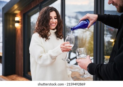 Cheerful Woman Holding Glass And Smiling While Man Pouring Champagne From Bottle. Couple In Love Enjoying Alcoholic Drink Outside Scandinavian House Barnhouse. Relationship And Celebration Concept.