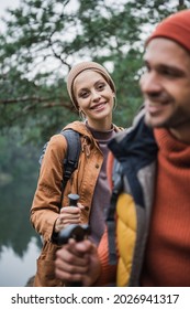 Cheerful Woman Holding Hiking Stick And Looking At Boyfriend On Blurred Foreground