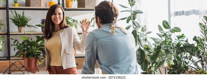 Cheerful Woman Holding Plant And Waving Hand At Florist In Flower Shop, Banner