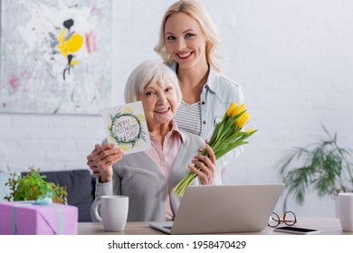 Cheerful Woman Hugging Mother With Happy Mothers Day Lettering On Greeting Card And Flowers Near Laptop