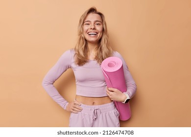 Cheerful woman leads sporty lifestyle dressed in activewear hold rolled mat being in good mood prepares for training in gym smiles broadly isolated over brown background. Regular workout concept - Shutterstock ID 2126474708