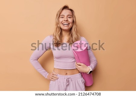 Cheerful woman leads sporty lifestyle dressed in activewear hold rolled mat being in good mood prepares for training in gym smiles broadly isolated over brown background. Regular workout concept
