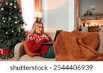 Cheerful woman lies on a couch in a beautiful living room by the Christmas tree.Wrapped in a blanket and wearing a red sweater with a festive headband, she works on laptop in a cozy atmosphere.