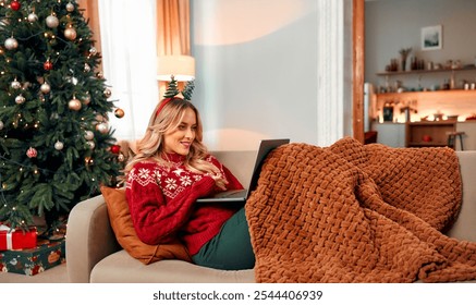 Cheerful woman lies on a couch in a beautiful living room by the Christmas tree.Wrapped in a blanket and wearing a red sweater with a festive headband, she works on laptop in a cozy atmosphere. - Powered by Shutterstock