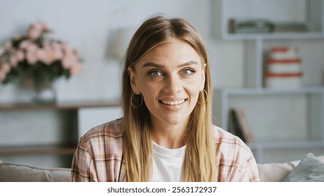 Cheerful woman with long blond hair and gold hoop earrings smiling brightly in a cozy living room adorned with a bookshelf and a vase of flowers - Powered by Shutterstock