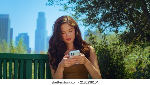 Cheerful Woman Looking Back On Blurred Cityscape Holding Smartphone In Green Park Close Up. Attractive Asian Girl Use Modern Gadget Sitting On Bench Outdoors. Cute Lady Smiling Looking On Phone Screen