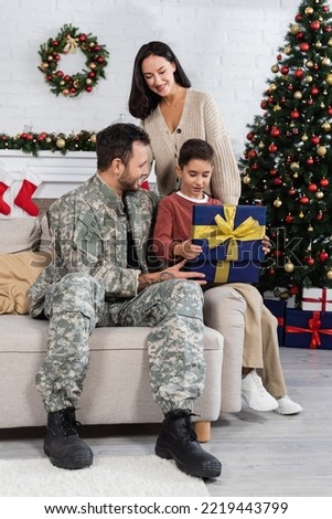 cheerful woman looking at son opening gift box near father in camouflage and decorated christmas tree