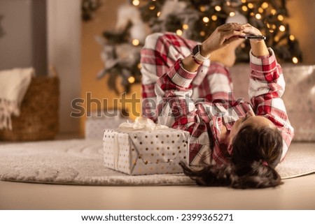 Cheerful woman in pajamas lying on carpet in living room with gift next to Christmas tree doing video chat with friends.