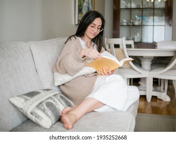 Cheerful Woman Reading A Yellow Book On The Coach Of Her Home. Woman Laughing At A Funny Story Enjoying Spending Time At Home. Concept Spending Time At Home.