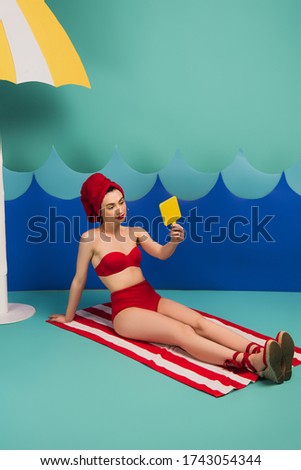 cheerful woman in red towel and swimsuit looking at mirror near blue paper cut waves