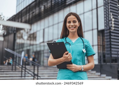 A cheerful woman in scrubs holds a clipboard, standing confidently before a modern hospital building, representing professional healthcare service. - Powered by Shutterstock