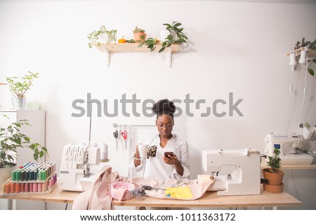 Cheerful woman seamstress in apron talking on phone and drinking coffee