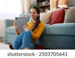 A cheerful woman sits comfortably on the floor, tablet in hand, engaged in a videocall. She leans against her couch in a cozy living room, sipping coffee during her relaxed conversation.