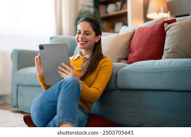 A cheerful woman sits comfortably on the floor, tablet in hand, engaged in a videocall. She leans against her couch in a cozy living room, sipping coffee during her relaxed conversation. - Powered by Shutterstock