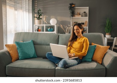 A cheerful woman is sitting at her cozy apartment and using a laptop for browsing and searching online on the internet. A young woman is sitting on sofa in her modern apartment and smiling at laptop. - Powered by Shutterstock