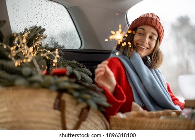 A Cheerful Woman Smiling And Looking At Sparkler While Sitting In Car Trunk , Traveling By Car During Christmas Holidays, Have A Holiday Mood. High Quality Photo