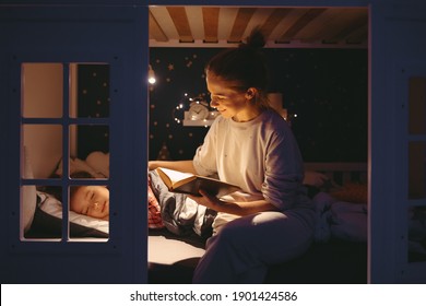 Cheerful woman smiling and reading fairytale to sleeping girl while sitting on illuminated bed in cozy bedroom at night at home - Shutterstock ID 1901424586