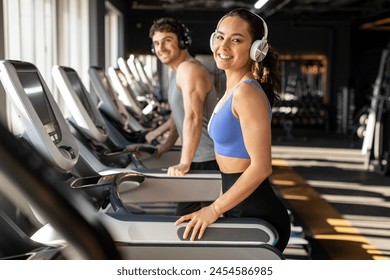 Cheerful woman in sportwear and headphones exercise running treadmill in fitness gym health club, smiling at camera. Calories burn during cardio training - Powered by Shutterstock