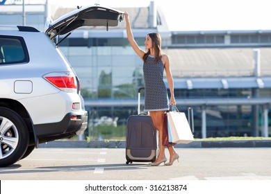 Cheerful woman is standing near her car and closing the trunk. She is holding many packets of bought things and smiling. There is a suitcase near her. Copy space in right side