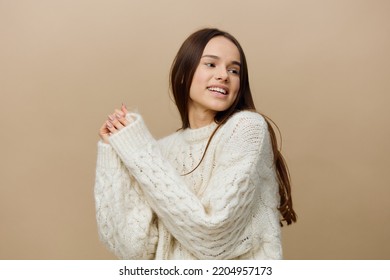 a cheerful woman is standing sideways on a light brown background in a white sweater, looking excitedly to the side with her hands raised in front of her