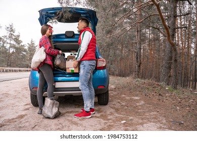 Cheerful Woman In Sunglasses Holding Trash Bag While Man Standing With Carton Box Near Car In Forest 