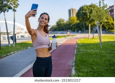 A cheerful woman takes a selfie while jogging on a sunny day, showcasing fitness and healthy lifestyle. She holds a water bottle, wearing sportswear, on a scenic outdoor path. - Powered by Shutterstock