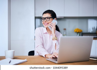 Cheerful woman talking on phone while working on laptop from home