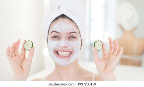 Cheerful Woman With A Towel On Her Hair And In A Clay Face Mask Fooling Around With Cucumbers In Her Hands. Taking Care Of Beauty At Home