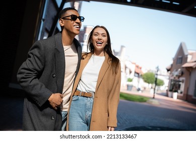 cheerful woman in trendy coat laughing near african american man in sunglasses on street in city - Shutterstock ID 2213133467