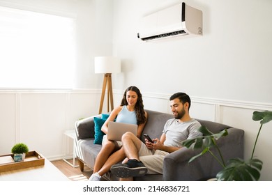 Cheerful woman using the laptop and young man texting while relaxing together at home with ac unit on