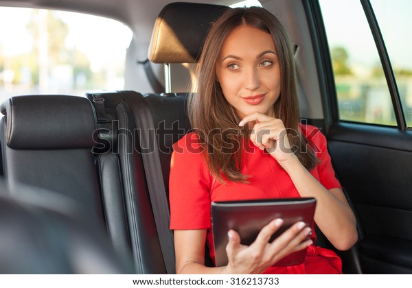 Cheerful woman is using a tablet in\
car. She is sitting on back seat and smiling. The lady is\

