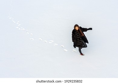 A Cheerful Woman In A Warm Fur Coat Walks Across The Open Field Leaving Footprint In The Snow