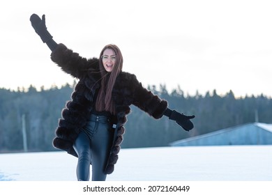 A Cheerful Woman In A Warm Fur Coat Walks Across The Open Field Leaving Footprint In The Snow