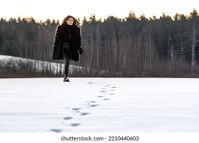 A Cheerful Woman In A Warm Fur Coat Walks Across The Open Field Leaving Footprint In The Snow