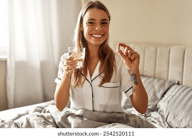 A cheerful woman wearing comfy pajamas happily beams as she holds a glass of refreshing water and a nutritional supplement, enjoying her fulfilling morning routine in her cozy, inviting bedroom space - Powered by Shutterstock