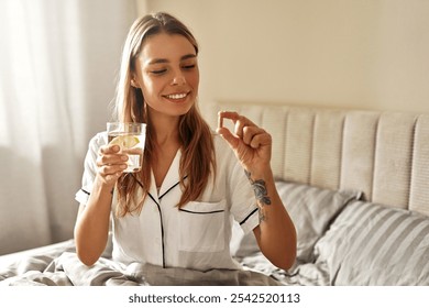 A cheerful woman wearing comfy pajamas happily beams as she holds a glass of refreshing water and a nutritional supplement, enjoying her fulfilling morning routine in her cozy, inviting bedroom space - Powered by Shutterstock