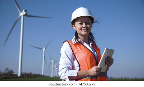 Cheerful Woman Wearing Hard Hat Standing Against Turbines At Wind Farm On Sunny Day