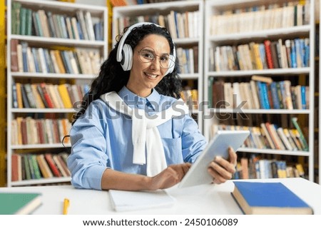 A cheerful woman wearing headphones is comfortably using a digital tablet in a well-stocked library, surrounded by numerous books.