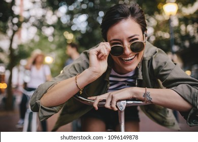 Cheerful woman wearing sunglasses leaning on her bike outdoors. Woman enjoying outdoors with friends at the back. - Powered by Shutterstock