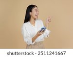 A cheerful woman in a white blouse smiling and pointing sideways while using her smartphone, conveying excitement and engagement against a beige background