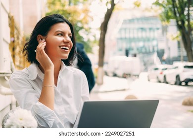 Cheerful woman is working at the laptop outdoors at a cafe table on the street and wearing wireless ear pods.