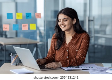 A cheerful woman, works on her laptop in a modern office. The setting features colorful sticky notes on glass and paperwork on the desk, highlighting productivity and professional focus. - Powered by Shutterstock