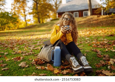 Cheerful woman in a yellow jacket sits among colorful autumn leaves, pouring a hot beverage into a cup while enjoying a peaceful moment outdoors. Autumn landscape. - Powered by Shutterstock