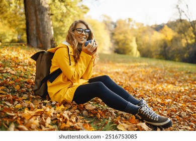 Cheerful woman in a yellow jacket sits among colorful autumn leaves, pouring a hot beverage into a cup while enjoying a peaceful moment outdoors. Autumn landscape. - Powered by Shutterstock