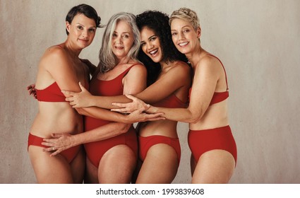 Cheerful women of different ages embracing their natural and aging bodies. Four happy and body positive women embracing each other while wearing red underwear against a studio background.