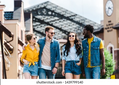 cheerful women with hands in pockets walking near multicultural men - Shutterstock ID 1439799140