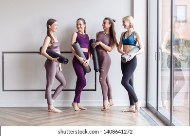 Cheerful Women Wearing Activewear Standing Near Large Window Hold Yoga Mats Ready Start Fitness Training And Rejoice At Meeting. The Concept Of Sports Lifestyle And Like-minded People. Yoga Concept