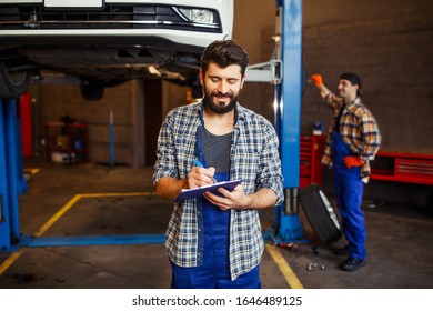 Cheerful Worker In Uniform Taking Notes And Automechanic Behind Woking Together