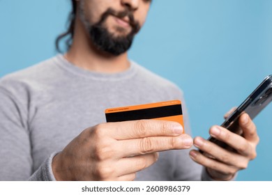 Cheerful young adult does shopping online with his smartphone and card, entering his banking credentials on a website. Person illustrating e commerce, digital payment and modern habits. Close up.